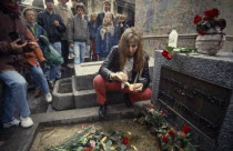 Pere Lachaise Cemetery. Visitors surrounding Jim Morrison grave with a woman knelling by the grave stone lighting a candleJames