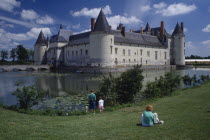 People sat on the bank of the moat looking towards the chateau.