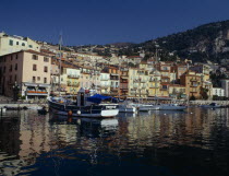 Villefranche sur Mer  harbour with moored fishing boats and yachts  colourful quayside buildings and houses on hillside behind.