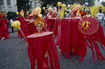 Notting Hill  Carnival. Little girl in red costume and flame shaped head-dress  adults in costume blowing whistles behind her.