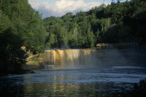 Tahquamenon Falls waterfall surrounded by lush greenery and man standing on riverbank nearby