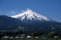 Pucon.  Villarica Volcano 2847m.  Active volcano in the Chilean Andes  snow covered cone rising above forest covered hills with building rooftops in the foreground.