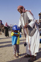 Man with young child jockey dressed in racing silks at camel racing event.