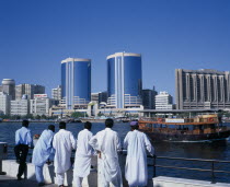 Dubai Creek.  Men standing on waterfront looking out towards city skyline and passing dhow.Dubayy United Arab Emirates