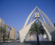 Dubai Clock Tower.  Modern sculptural structure with pool and fountains below and palm tree in foreground.  Traffic and city buildings behind.Dubayy United Arab Emirates