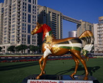 Brightly painted statue of horse with advertising for Dubai Real Estate Centre set in formal grounds with hotel and high rise buildings behind.  Center Dubayy United Arab Emirates