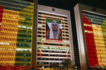 Portrait of the former Amir HH Sheikh Sabah Al-Ahmad Al-Jaber Al-Sabah flanked by Kuwait flag made from strings of illuminated lights on building at night.Emir