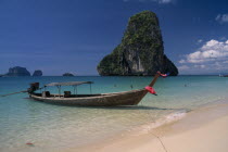 Longtail boat moored in shallow water beside golden sand beach with people swimming and limestone karst rock formation behind.