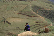 Hmong woman sitting on hillside overlooking agricultural terraces.