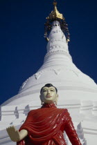 White stupa at newly painted temple on road to Mandalay with standing Buddha in pose with extended right arm with open palm turned to front.Pose is possibly  Charity  where theBuddha is either grant...