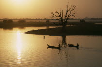 View from U Bein Bridge at Amarapura near Mandalay with canoes silhouetted against water reflecting golden sunset and trees and landscape in drifts of mist. Burma