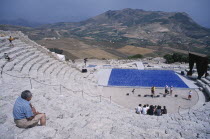 Graeco Roman Amphitheatre. Man sat on steps looking down over stage area with a group of people sat on the steps at the bottom having their photograph taken near stage lighting equipment