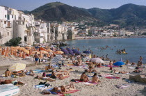 Cefalu. View across busy sandy beach with sunbathers on sand and swimming in the sea overlooked by waterfront apartments and hills