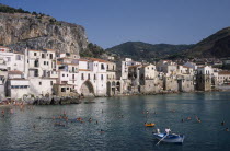 Cefalu. View across people swimming in the sea next to sandy beach with a man rowing a small boat in the foreground overlooked by waterfront apartments and hills behind
