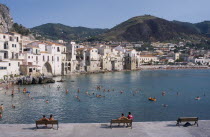 Cefalu. View from stone walls with people sat on benches towards sunbathers swimming in the sea overlooked by waterfront apartments and hills behind