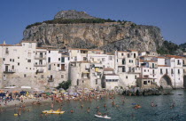 Cefalu. View across the sea with people swimming in the water and sunbathers on busy sandy stretch of beach towards waterfront apartments overlooked by hills behind