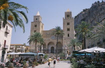 Cefalu. Pathway towards II Duomo Norman Cathedral between cafes with people sat at table and chairs near palm trees