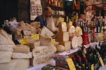 Market cheese stall with a male stall holder displaying a selection of cheeses and olive oil with euro money price signs