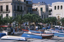 Mondello. Fishing boats moored next to the promenade overlooked by buildings with people walking along path near palm trees