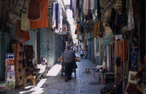 An Arab man pushing his bread cart through a narrow street between clothes and textile stalls in the Old City