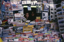Two women looking out from newsagents stall completely surrounded by newspapers and magazines.