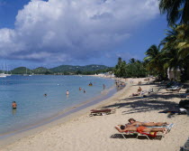 Sunbathers and swimmers on sandy beach lined with palm trees. Yachts seen on the water and green hills behind