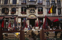 Grand Place.  Busy cafe with people sitting at outside tables in the sunshine.  Part view of building facade with stone balcony and flower filled window boxes.UNESCO World Heritage Site  UNESCO Worl...