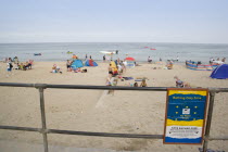 View through a railing with a Beach Safety sign towards busy sandy beach with sunbathers on the sandGreat Britain United Kingdom UK