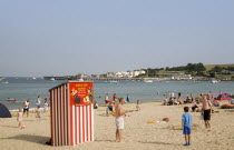 Small red and white striped Punch and Judy theatre on sandy beach with sunbathers on the sand and swimming in the sea.Great Britain United Kingdom Theater UK