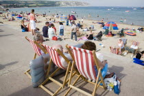 People sitting on red and white striped deckchairs looking out towards busy sandy beach with sunbathers on the sandGreat Britain United Kingdom UK