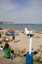 View over busy sandy beach with sunbathers on the sand towards the sea. Viewing telescope in the foreground.Great Britain United Kingdom UK