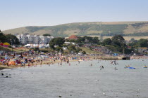 View across the sea towards the shoreline of sandy beach with sunbathers on the sand and swimming in the sea. Hotel buildings and hills behind.Great Britain United Kingdom UK