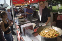 French Market. Stallholder cooking chicken and vegetable dishes in large woks with a customer waitingGreat Britain United Kingdom UK