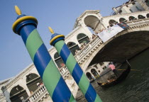 Colourful posts for mooring boats in front of gondolas carrying sightseers on the Grand Canal beneath the Rialto Bridge lined with tourists Colorful