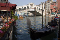 A gondola moored beside a restaurant on the Grand Canal with tourists dining. Beyond the Rialto Bridge lined with tourists spans the canal