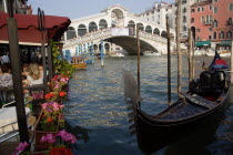 A gondola moored beside a restaurant on the Grand Canal with tourists dining. Beyond the Rialto Bridge lined with tourists spans the canal