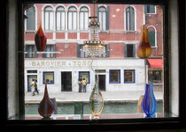 Tourists walking along the Fondamenta dei Vetrai looking in the windows of a glass shop gallery beside the Rio dei Vetrai canal on the lagoon island of Murano. Seen through a window display of modern...