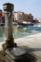 A free public drinking water fountain on the Canale di San Donato on the island of Murano. One of many on the island. A lagoon barge passes down the canal