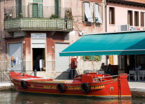 A traditional red lagoon barge moored alongside the Fondamenta Sebastiano Santi beside a grocery shop and coffee shop by the Ponte de la Terese bridge across the Canale di San Donato on the island of...