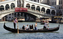 A Traghetto carrying local people on board crosses the Grand Canal. Tourists crowd on the Rialto Bridge spanning the canal with vaporetto passing beneath