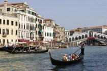 A Traghetto with local people on board crosses the Grand Canal and a Gondola carries tourists down river. Tourists crowd on the Rialto Bridge spanning the canal