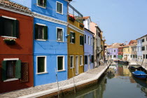 Colourful houses beside a canal on the lagoon island of Burano with tourists walking past boats moored alongside the edge of the canal. Colorful