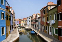 Colourful houses beside a canal on the lagoon island of Burano with tourists walking past boats moored alongside the edge of the canal. Colorful