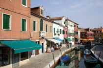 Colourful houses beside a canal on the lagoon island of Burano with boats moored alongside the edge of the canal. Tourists sit at tables outside a restaurant whilst others walk past shops by a bridge....