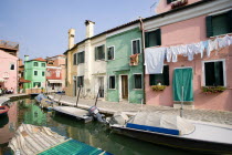 Colourful houses beside a canal on the lagoon island of Burano with boats moored alongside the edge of the canal. Tourists sit at tables outside a restaurant whilst others walk past shops by a bridge....