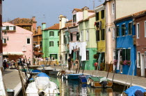 Colourful houses beside a canal on the lagoon island of Burano with people walking past boats moored alongside the edge of the canal. Tourists sit at tables outside a restaurant whilst others walk pas...