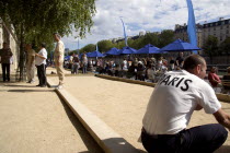 The Paris Plage urban beach. A man wearing a Paris t-shirt beside people playing boules petanque along the Voie Georges Pompidou a usually busy road closed to traffic opposite the Ile de la Cite