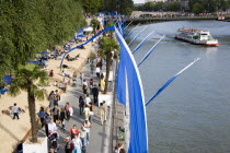 The Paris Plage urban beach. People strolling between the River Seine and other people lying on sand along the Voie Georges Pompidou usually a busy road now closed to traffic. A Pleasure boat on the r...