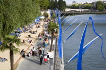 The Paris Plage urban beach. People strolling between the River Seine and other people lying on sand along the Voie Georges Pompidou usually a busy road now closed to traffic