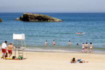 The Basque seaside resort on the Atlantic coast. Lifeguards and young tourists on the Grande Plage beach with a man paddling on a surfboard off the beach